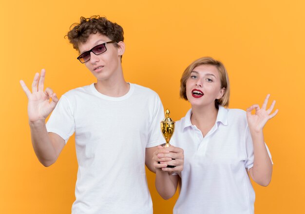 Jovem casal desportivo, homem e mulher, juntos segurando um troféu, mostrando uma placa de ok, sorrindo em pé sobre a parede laranja