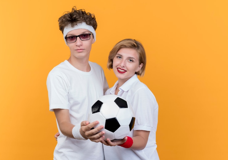 Foto grátis jovem casal desportivo, homem e mulher, juntos segurando uma bola de futebol, sorrindo por cima da parede laranja