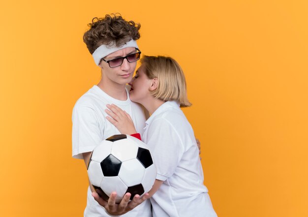 Jovem casal desportivo mulher triste a abraçar um homem enquanto ele segura uma bola de futebol em pé sobre a parede laranja