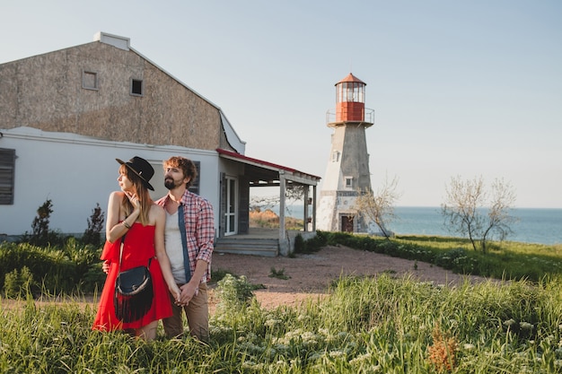 Jovem casal elegante apaixonado no campo, estilo indie hippie boêmio, férias de fim de semana, roupa de verão, vestido vermelho, grama verde, de mãos dadas, sorrindo