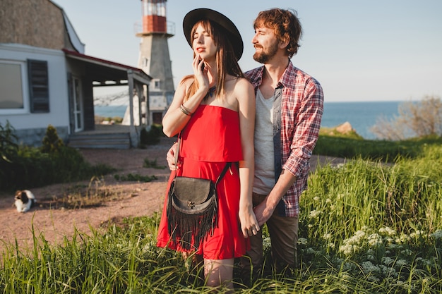 Jovem casal elegante apaixonado no campo, estilo indie hippie boêmio, férias de fim de semana, roupa de verão, vestido vermelho, grama verde, de mãos dadas, sorrindo