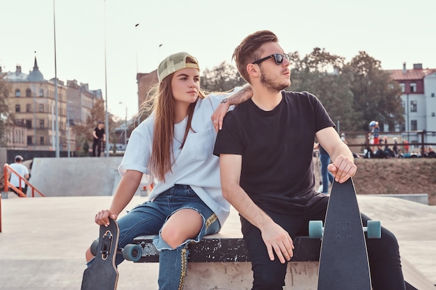 Jovem casal elegante de skatistas vestidos na moda relaxando no skatepark.