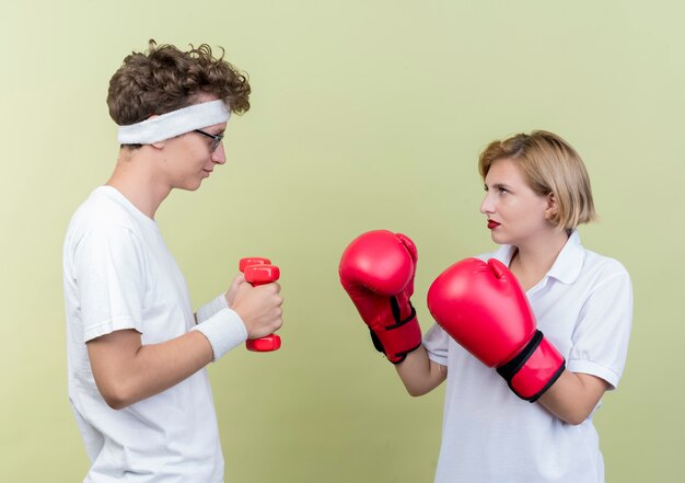 Jovem casal esportivo homem com halteres olhando para a namorada com luvas de boxe sorrindo sobre a luz