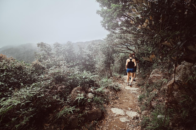 Foto grátis jovem casal está correndo pelo caminho nas montanhas florestais. há matagais verdes e árvores ao redor. nevoeiro e nuvens ao fundo.