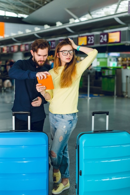 Jovem casal está parado no aeroporto com duas malas perto. ela tem cabelo comprido, suéter, jeans e tablet na mão. ele usa barba, camisa preta e calça. eles parecem um pouco chateados, talvez perdidos.
