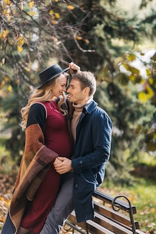 Jovem casal feliz, caucasiano, posando ao ar livre no dia de outono, uma jovem grávida abraça seu homem e ...