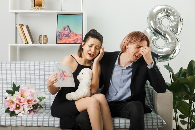 Jovem casal feliz dia da mulher com ursinho de pelúcia e cartão postal sentado no sofá da sala