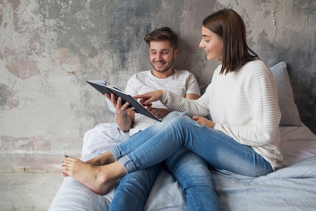 Jovem casal feliz e sorridente, sentado na cama em casa, com roupa casual, lendo um livro de jeans, um homem e uma mulher passando um tempo romântico juntos
