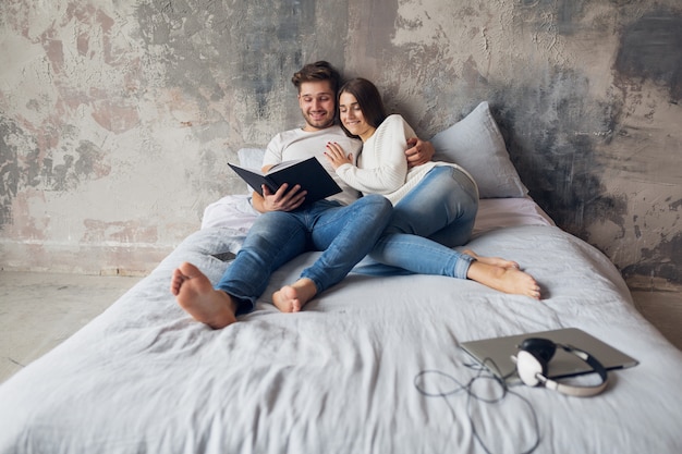Jovem casal feliz e sorridente, sentado na cama em casa com roupa casual, lendo um livro de jeans, um homem e uma mulher passando um tempo romântico juntos