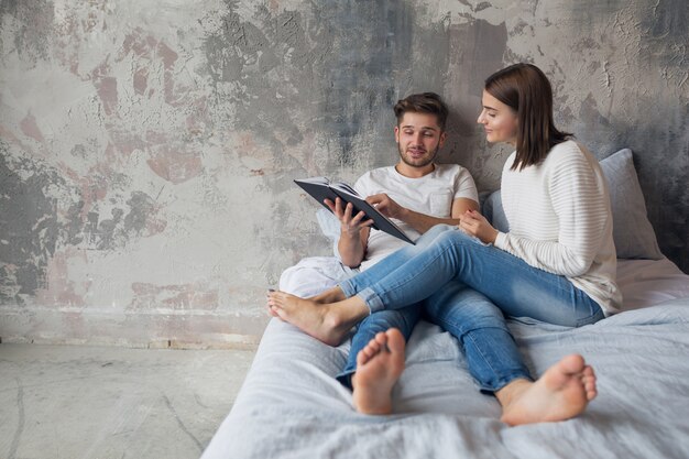 Jovem casal feliz e sorridente, sentado na cama em casa com roupa casual, lendo um livro de jeans, um homem e uma mulher passando um tempo romântico juntos