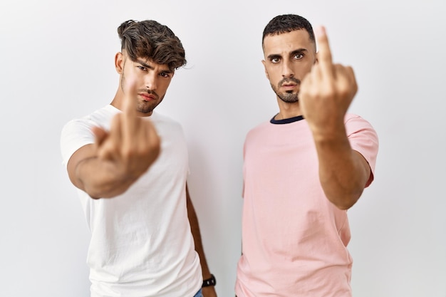 Jovem casal gay em pé sobre fundo isolado mostrando dedo médio, expressão indelicada e rude foda-se