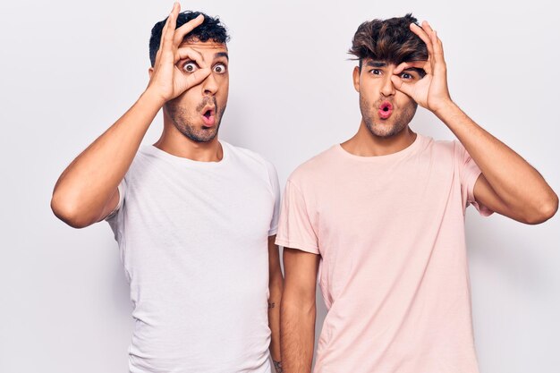Jovem casal gay vestindo roupas casuais fazendo gesto ok chocado com cara de surpresa, olho olhando por entre os dedos. expressão incrédula.