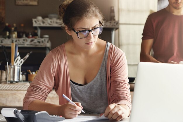 Jovem casal gerenciar o orçamento familiar. revisando suas contas bancárias usando notebook e calculadora na cozinha. Mulher atraente, concentrado olhar pagando impostos on-line no laptop pc