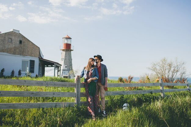 Jovem casal hippie estilo indie apaixonado caminhando pelo campo, de mãos dadas, farol no fundo, dia quente de verão, ensolarado, roupa boêmia, chapéu