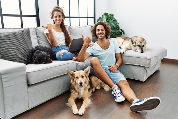 Jovem casal hispânico com cães relaxando em casa apontando para trás com a mão e polegares para cima, sorrindo confiante