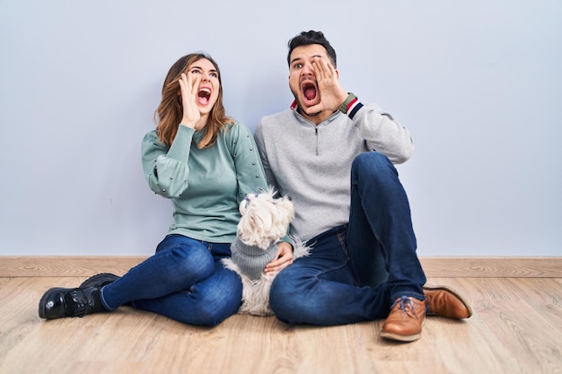 Jovem casal hispânico sentado no chão com cachorro gritando e gritando alto a lado com a mão no conceito de comunicação da boca