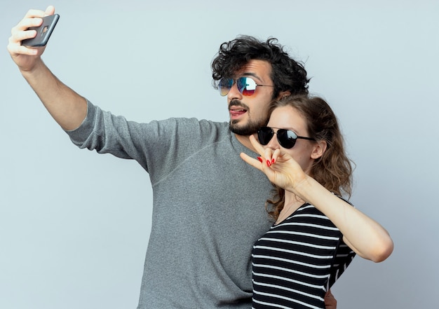 Foto grátis jovem casal homem e mulher felizes no amor, homem feliz tirando fotos deles usando o smartphone em pé sobre uma parede branca