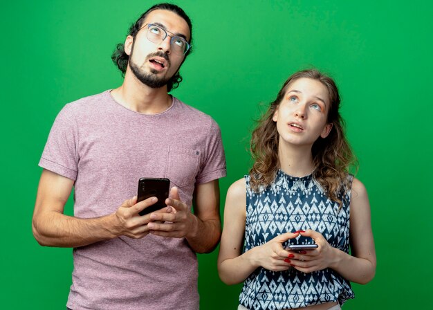 jovem casal, homem e mulher, olhando perplexo enquanto segura smartphones em pé sobre uma parede verde