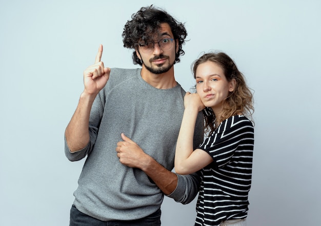 Foto grátis jovem casal, homem e mulher, sentando-se lado a lado, homem mostrando o dedo indicador, enquanto a namorada franzindo a testa em pé sobre uma parede branca
