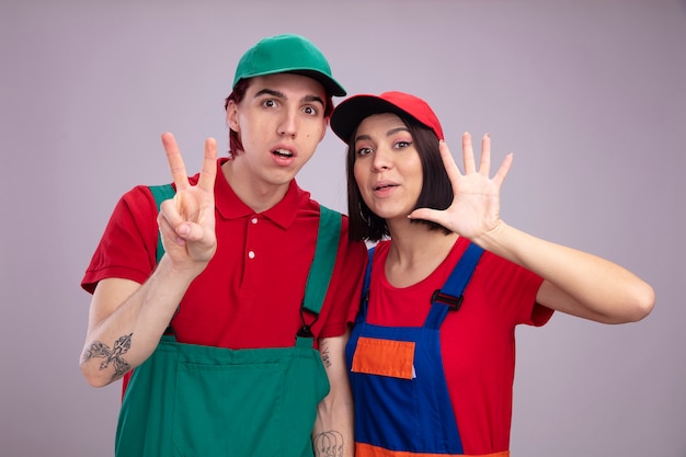 Jovem casal impressionado com uniforme de trabalhador da construção civil e boné olhando para o cara da câmera mostrando dois com a mão garota mostrando cinco com a mão isolada na parede branca