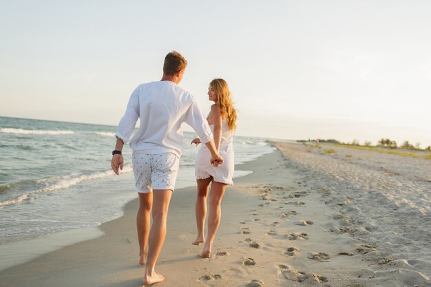 Jovem casal lindo caminha à beira-mar ao pôr do sol.