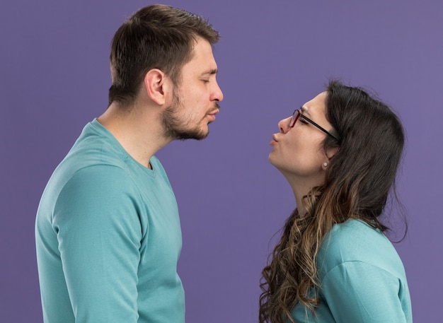 Jovem casal lindo com roupas casuais azuis, homem e mulher vão se beijar, felizes e apaixonados em pé sobre a parede roxa