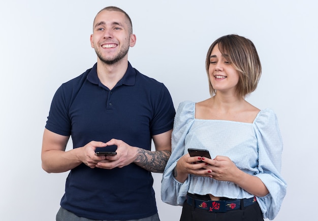 Jovem casal lindo com roupas casuais, homem e mulher feliz e positiva segurando smartphones, olhando de lado com um sorriso no rosto em pé