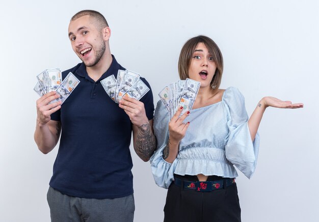 Jovem casal lindo com roupas casuais, homem e mulher segurando dinheiro, sorrindo alegremente em pé sobre uma parede branca