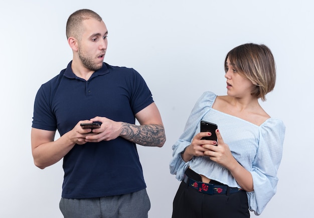 Foto grátis jovem casal lindo com roupas casuais, homem e mulher segurando smartphones, olhando um para o outro confuso e muito ansioso em pé