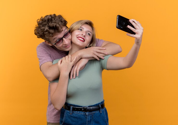 Foto grátis jovem casal lindo dançando junto, usando um celular, tirando uma selfie e sorrindo sobre a parede laranja