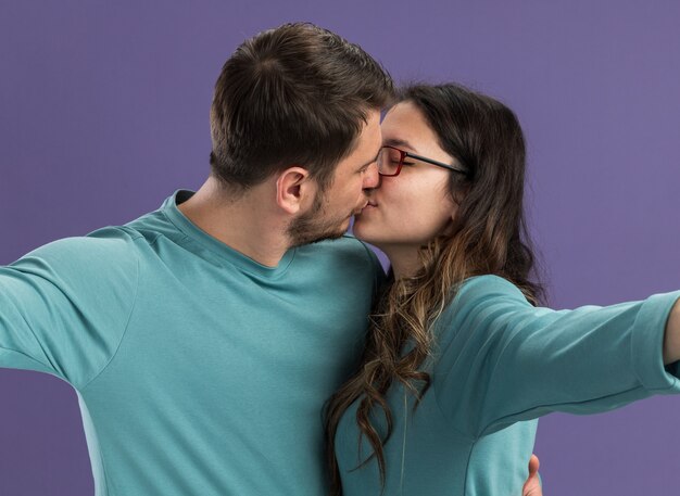 Jovem casal lindo em roupas casuais azuis, homem e mulher se beijando, felizes e apaixonados, em pé sobre a parede roxa
