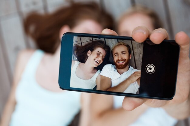 Jovem casal lindo fazendo selfie, sorrindo, deitado sobre tábuas de madeira, mostrando a tela do telefone