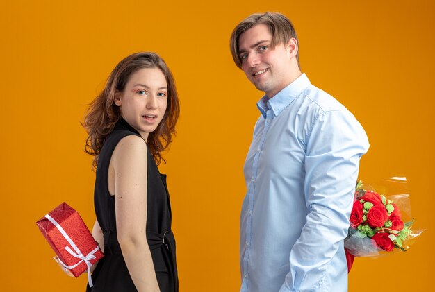 jovem casal lindo feliz homem com um buquê de rosas vermelhas nas costas e uma mulher com um presente olhando para a câmera sorrindo alegremente comemorando o dia dos namorados em pé sobre um fundo laranja