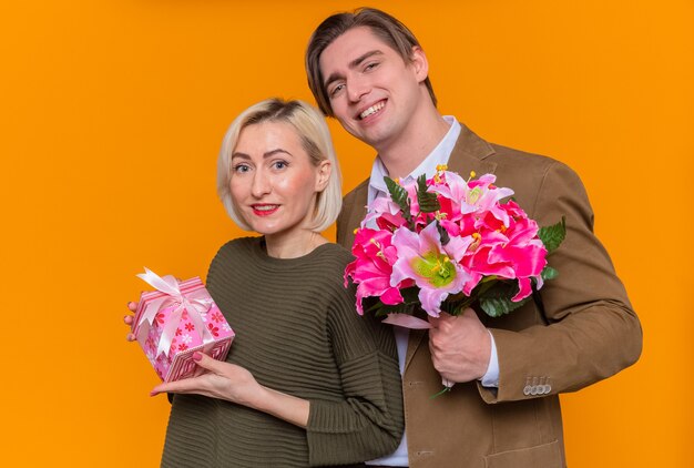 Jovem casal lindo feliz segurando um buquê de flores e uma mulher com um presente feliz e apaixonado, celebrando o dia internacional da mulher em pé sobre a parede laranja