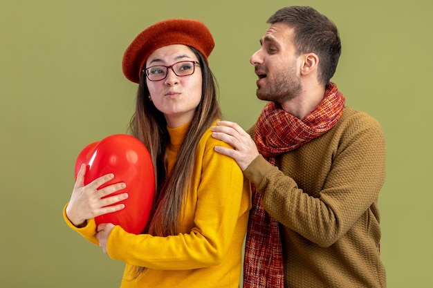 jovem casal lindo feliz tocando sua namorada confusa na boina com um balão em forma de coração comemorando o dia dos namorados em pé sobre a parede verde