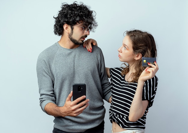 Jovem casal lindo homem e mulher confuso, segurando o smartphone e olhando para a namorada dela, que está segurando o cartão de crédito dele sobre fundo branco