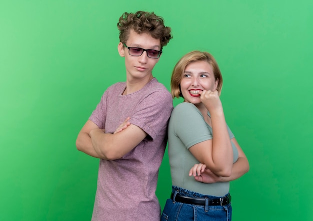 Jovem casal lindo homem e mulher de pé, costas contra costas, felizes e sorrindo positivamente sobre a parede verde