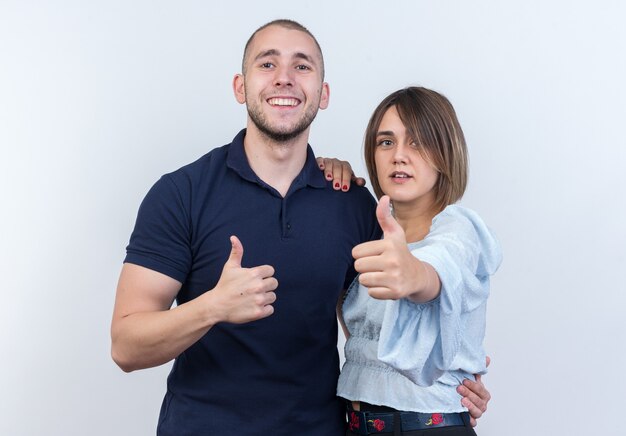 Jovem casal lindo homem e mulher felizes e positivos sorrindo alegremente mostrando os polegares em pé sobre uma parede branca