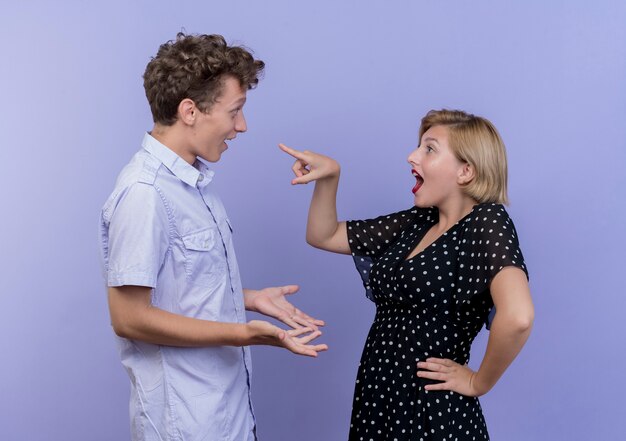 Jovem casal lindo homem e mulher mulher feliz e positiva apontando com o dedo para o namorado surpreso em pé sobre a parede azul