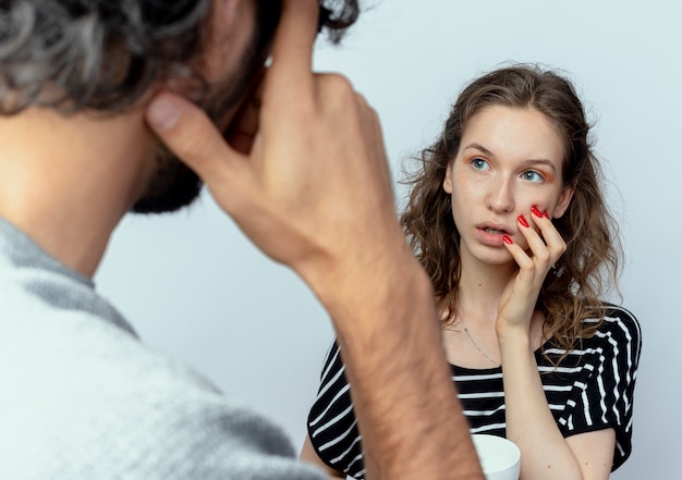 Jovem casal lindo, homem e mulher, olhando um para o outro, confusos sobre um fundo branco