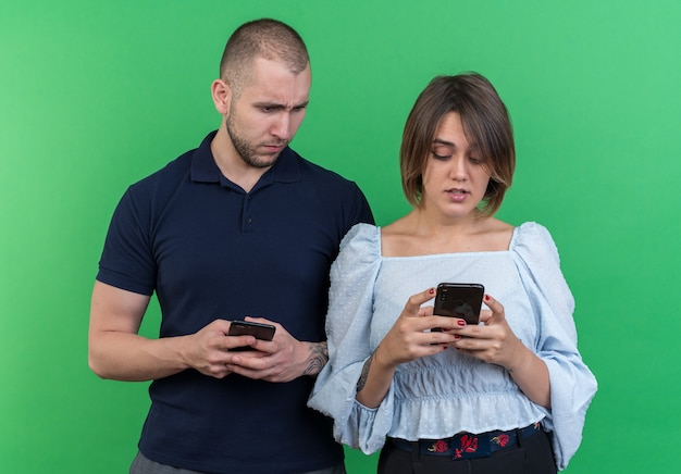 Jovem casal lindo homem e mulher segurando smartphones homem olhando desconfiado para o smartphone da namorada dele em pé