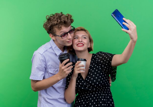 Jovem casal lindo juntos segurando xícaras de café, usando telefone celular, tirando selfie e sorrindo sobre a parede verde