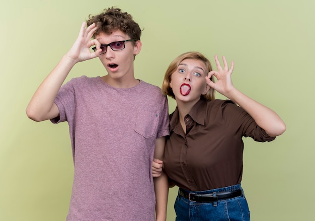 Jovem casal lindo menino e menina vestindo roupas casuais se divertindo, sorrindo, mostrando a língua para fora, mostrando sinal de ok sobre a luz