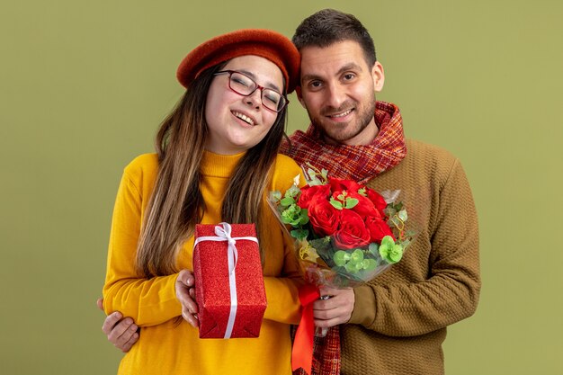 jovem casal lindo mulher feliz em boina com presente e homem com buquê de rosas vermelhas, olhando para a câmera, sorrindo, feliz e apaixonado juntos, comemorando o dia dos namorados em pé sobre a parede verde