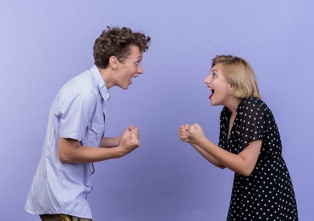 Foto grátis jovem casal lindo olhando um para o outro com os punhos cerrados, feliz e animado sobre a parede azul