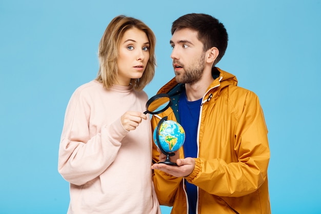 Jovem casal lindo posando sobre parede azul Homem vestindo casaco de chuva segurando o globo pequeno. Menina com lupa.