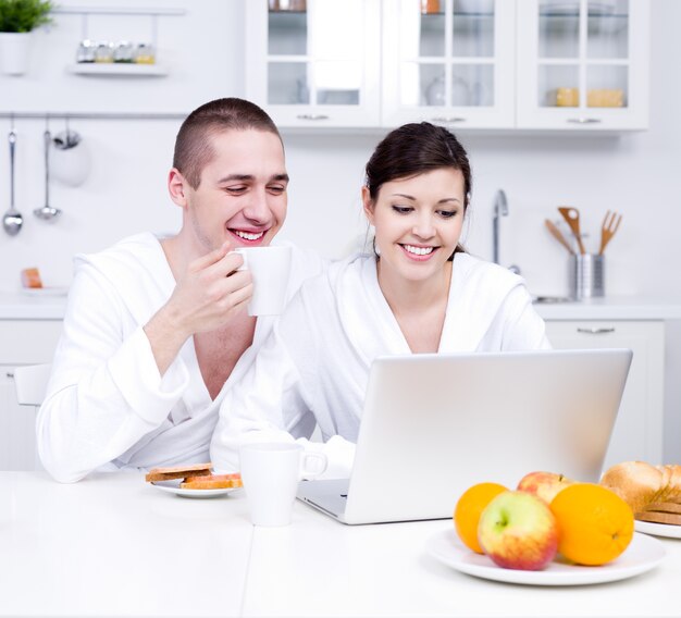 Jovem casal lindo sentado na cozinha olhando para o laptop