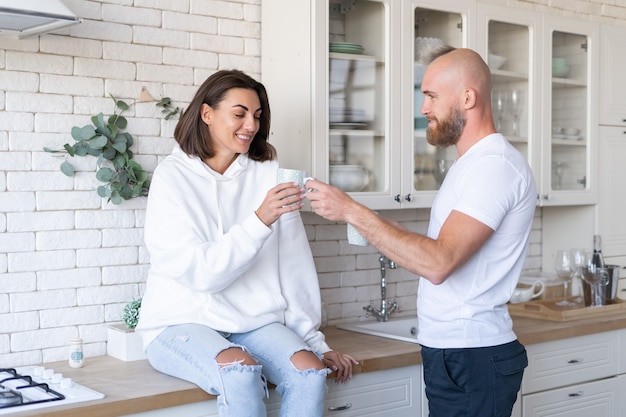 Jovem casal, marido e mulher em casa na cozinha, sorriso feliz, risada, tomam café pela manhã