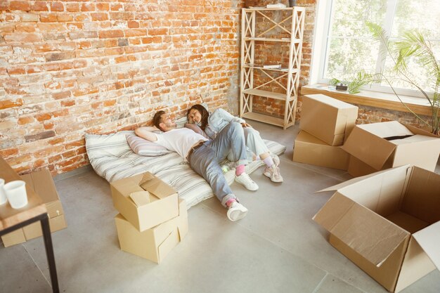 Jovem casal mudou-se para uma nova casa ou apartamento. Deitados juntos, relaxando após a limpeza e desfazendo as malas no dia da mudança. Pareça feliz, sonhador e confiante. Família, mudança, relações, primeiro conceito de casa.