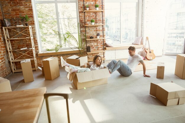 Jovem casal mudou-se para uma nova casa ou apartamento. Divertir-se com caixas de papelão, relaxar após limpar e desfazer as malas no dia da mudança